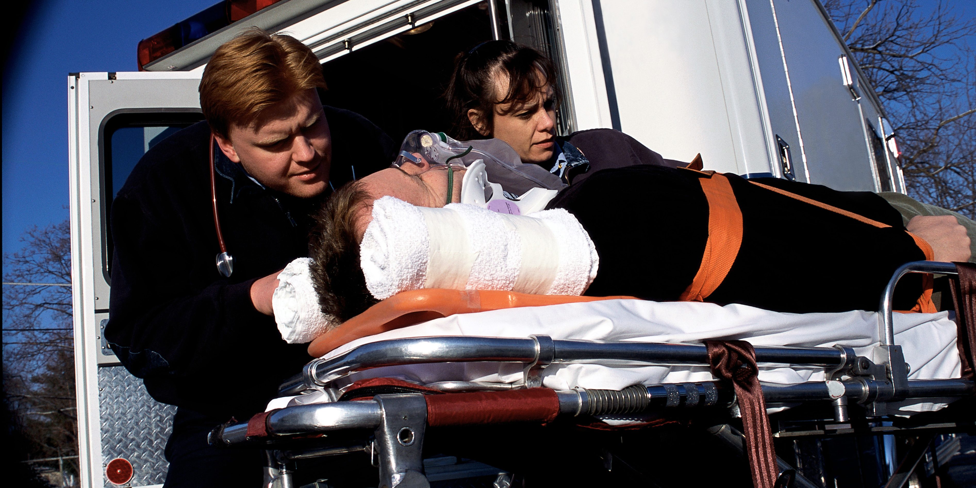 Two Caucasian paramedics, one male and one female, strapping an injured Caucasian male into a stretcher